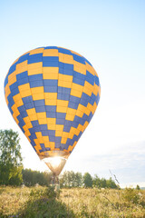 Obraz premium Hot air balloon with blue and yellow checkered pattern floating above grassy field during sunrise, basket carrying group of people enjoying outdoor adventure in rural landscape