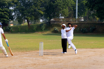 A cricketer in white delivers a ball with focus, while an umpire and batsman watch on a sunny cricket field