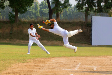 A wicketkeeper in white leaps to catch the ball as a fielder stands ready, on a cricket field