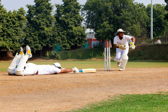 A wicketkeeper in white runs to stump the ball as a batsman dives to make the crease, on a grassy field