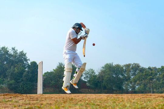 A cricketer in white jumps to avoid a fast-approaching ball on a grassy field, demonstrating agility and quick reflexes