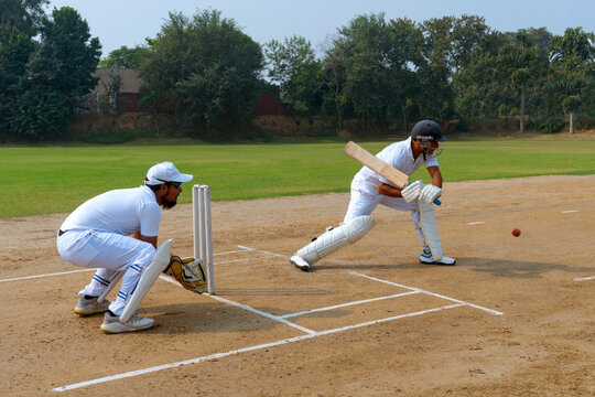 A cricketer in white uniform executing a shot with precision, while a wicketkeeper crouches behind on a field