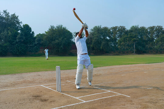 A cricketer in full swing, hitting the ball with force on a well-maintained field under a bright sky