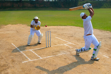 A batsman in white uniform hits a high shot, with a wicketkeeper ready behind the stumps on a cricket field