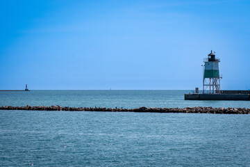 Fototapeta premium Serene blue seascape with a small white lighthouse on a stone breakwater, calm horizon and clear sky—perfect coastal scene for travel and photography.
