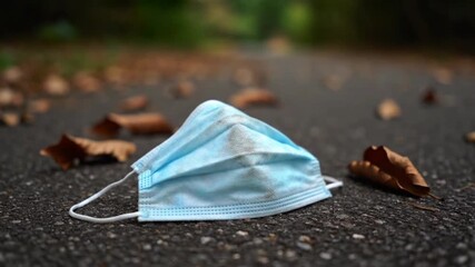 Dirty disposable blue medical face mask discarded on asphalt ground with fallen autumn leaves symbolizing environmental pollution and the lingering impact of th