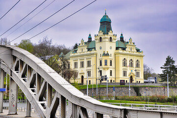 Ostrawa - Ratusz  Sląskiej Ostrawy (Radnice Slezské Ostravy). Okazały, żółty, zabytkowy budynek stoi obok metalowego mostu łukowego w spokojnej, miejskiej scenerii. © Pol Badger