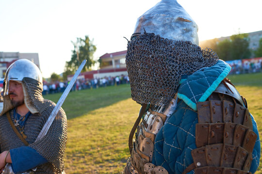 Two Caucasian men wearing medieval armor participating in historical reenactment outdoors, one holding sword while other standing in chainmail and plate armor, blurred crowd in background