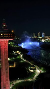 Aerial drone video pans left at night, revealing lit Skylon Tower, Niagara Falls city. Cascades glow with multi-colored mist. Travel documentary concept
