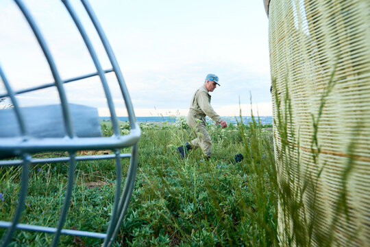 Caucasian man walking through grassy field near large wicker basket and metal structure, wearing uniform and cap, outdoor setting with distant landscape visible