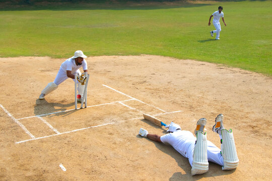 A batsman in white uniform dives to avoid a run-out, with a wicketkeeper ready behind the stumps on a cricket field