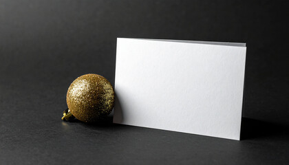 Plain white card standing against matte black background with single golden ornament.