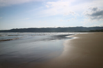 Obraz premium Calm, reflective water on a beach shoreline at low tide. Ocean, sky, and a distant hill line.