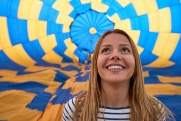 Portrait of Caucasian young adult woman smiling and looking upward while standing inside colorful hot air balloon envelope, geometric blue and yellow pattern visible overhead