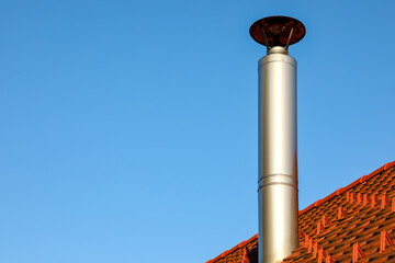  Metal chimney rises from a red tiled roof under a clear blue sky. Sunlight gleams on the polished flue capped with a round cover, symbolizing heating, ventilation, and modern architecture.