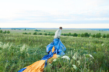 Young adult Caucasian man walking through grassy field dragging large blue and yellow parachute behind, facing away from camera, surrounded by wildflowers and open landscape