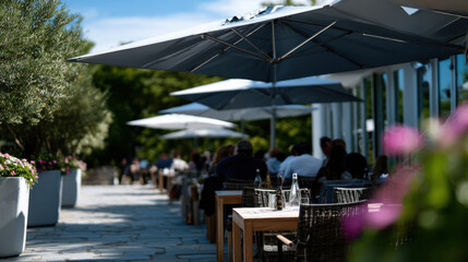Obraz premium A vibrant outdoor dining scene featuring sun shades, lush greenery, and patrons enjoying their meals in an inviting and bright setting under blue skies.