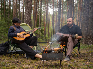 Young woman playing guitar while sitting near bonfire with man in forest during camping trip, creating cozy and relaxing atmosphere surrounded by nature