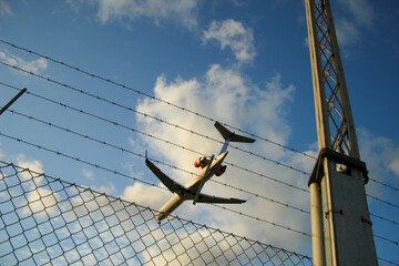 A passenger jet captured in flight, seen from below, framed by barbed wire and chain link fencing