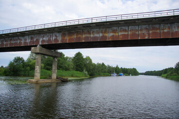 An old metal railway bridge with rusty girders crossing a calm river lined with green trees under a cloudy sky.