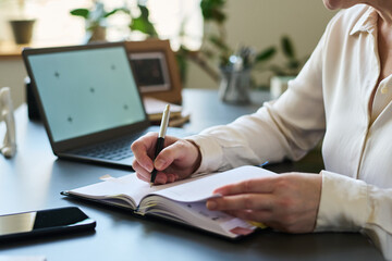 Middle aged woman writing notes in notebook while sitting at desk with open laptop and smartphone, hands visible, working in psychologist office, focusing on documentation