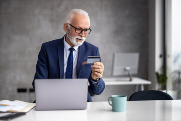 Senior professional checking card information while working on laptop in modern office setting during daytime