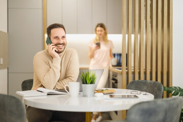 Smiling man on phone enjoying work from home while woman uses smartphone in modern kitchen