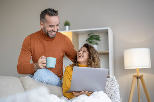 Couple sharing a joyful moment while enjoying coffee and working on a laptop in a cozy living room