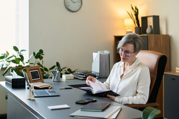 Middle aged woman sitting at desk reviewing documents with pen in hand, wearing eyeglasses, working in office setting with plants and shelves in background