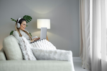 Woman using laptop while sitting on a cozy sofa with headphones in a well-lit living room
