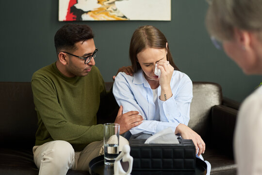 Woman crying and wiping tears while sitting on sofa, South Asian man comforting her, middle aged woman observing in foreground, psychologist session