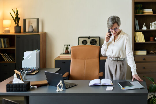Middle aged woman standing at desk talking on smartphone, reviewing open notebook and documents, modern office setting suggesting professional psychologist working