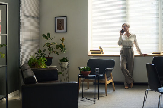 Middle aged woman standing near window talking on smartphone in psychologist office, indoor setting with plants and chairs visible, professional environment depicted