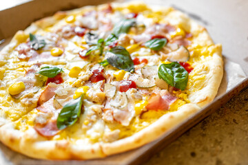 Thin crust pizza with ham, chicken, corn, basil, and melted cheese in a cardboard box. Freshly baked, golden brown, and garnished with black pepper.