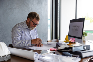 An older male architect or engineer reviews technical drawings at his desk, writing notes beside a computer screen displaying blueprints.