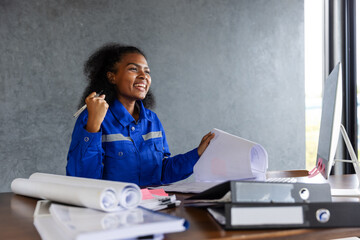 A smiling female engineer in a blue uniform reviews blueprints at her desk, surrounded by documents and a computer, symbolizing planning, design, and success in construction and engineering projects.