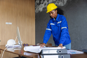 A female engineer in a blue uniform and yellow safety helmet reviews construction blueprints on a desk, symbolizing planning, architecture, and professional work in engineering and building projects.