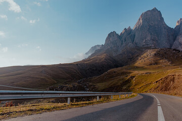 Winding road leads through a mountainous landscape. The scene features tall, rugged peaks under a blue sky with scattered clouds. Green hillsides are visible.