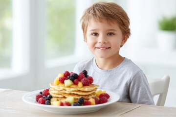 Young boy with light brown hair smiles joyfully at the camera, sitting at a table with a large plate of colorful pancakes topped with fresh fruits, showcasing a delightful breakfast scene
