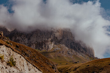 A rugged mountain landscape with steep cliffs and a cloudy sky. The terrain is rocky with patches of grass and shrubs.