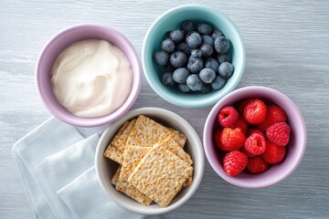 Colorful bowls filled with fresh blueberries, raspberries, creamy yogurt, and crispy crackers arranged on a light wooden surface, showcasing healthy snack options and vibrant colors