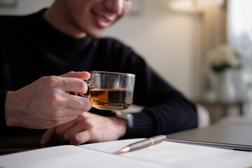 Close-up of a man holding a cup of tea with documents and a pen on the table.