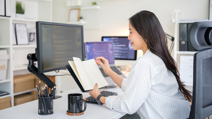 Woman developer writes in notebook while analyzing code on dual screens.