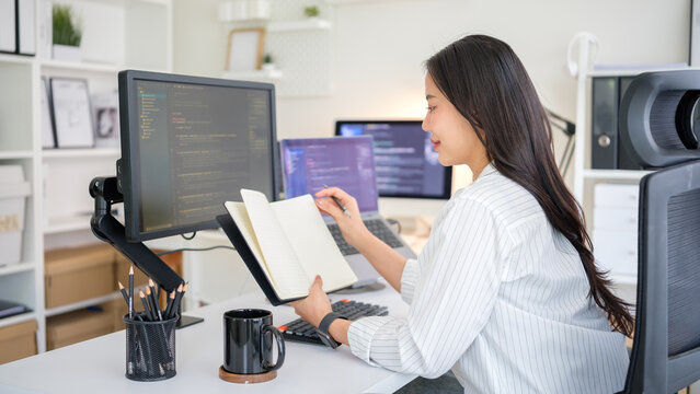 Woman developer writes in notebook while analyzing code on dual screens.