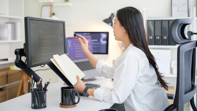 Female developer explaining code while pointing at monitor and holding a notebook.