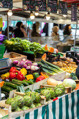 Fresh artichokes, peppers, and various other vegetables are on display in wooden crates at a street market. Unrecognizable people are in the background.