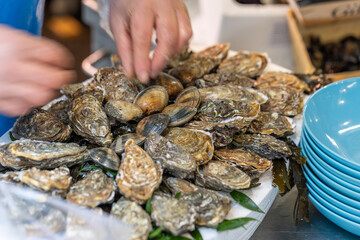 A vendor is arranging fresh oysters and clams on a white platter at a street seafood market in Paris, France. A stack of blue bowls is in the foreground.