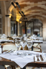 A set table with wine glasses, cutlery, and a napkin on a white tablecloth at an outdoor Parisian cafe. An arched brick ceiling is in the background.