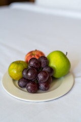 A plate of grapes, an orange,a and a red and green apple on white plate sitting on a bed at a resort