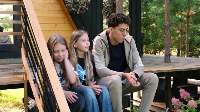 Loving interaction between brother and sisters sitting on wooden steps, laughter and communication in a serene environment, shared moments. Weekend family vacation at the cottage, communication
